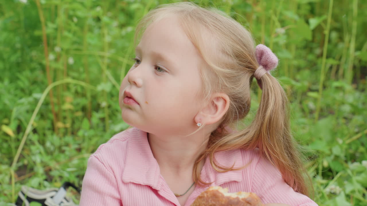 chica blanca sujetando una hogaza de pan en un prado, expresiones curiosas oliendo la corteza durante un picnic en un campo de hierba alta, suéter rosa, coletas, primer plano que transmite inocencia y slow living