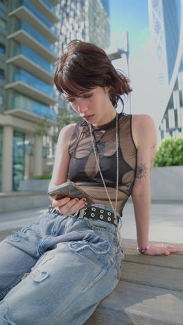 Vertical-Video-Outdoor-Fashion-Portrait-Of-Young-Alternative-Style-Woman-With-Tattoos-Sitting-On-Bench-At-Canary-Wharf-London-Messaging-On-Mobile-Phone-1