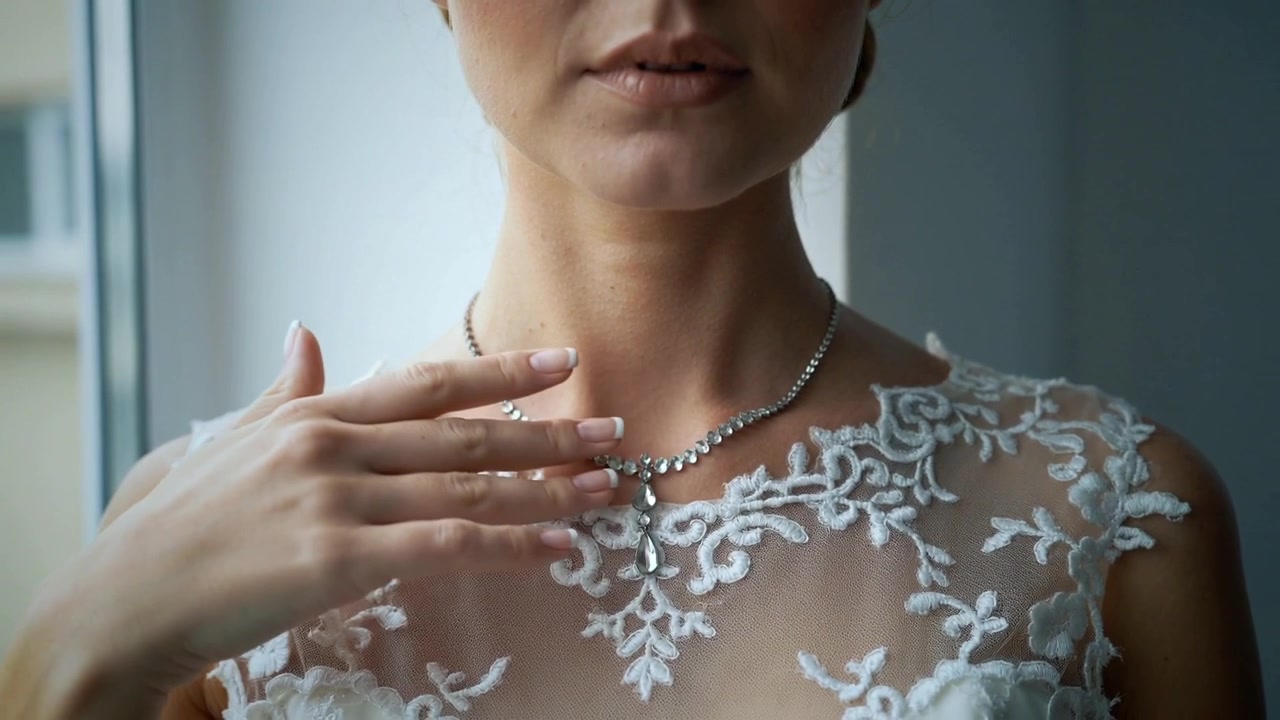 Donna in abito di pizzo bianco che, notando il ciondolo decentrato, sistema una collana di perline vicino alla finestra.