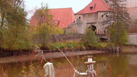 drone flies low over the lake and passes on an artificial little island with figures and a house and in front is the city gate of the old town and some roofs of houses