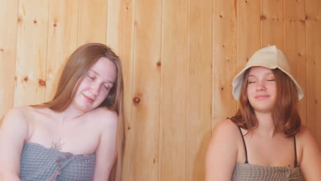 two women in towels smiling at each other in a cozy sauna, sharing a moment of happiness and relaxation, they are seated on a wooden bench with a serene wooden backdrop, enjoying their time