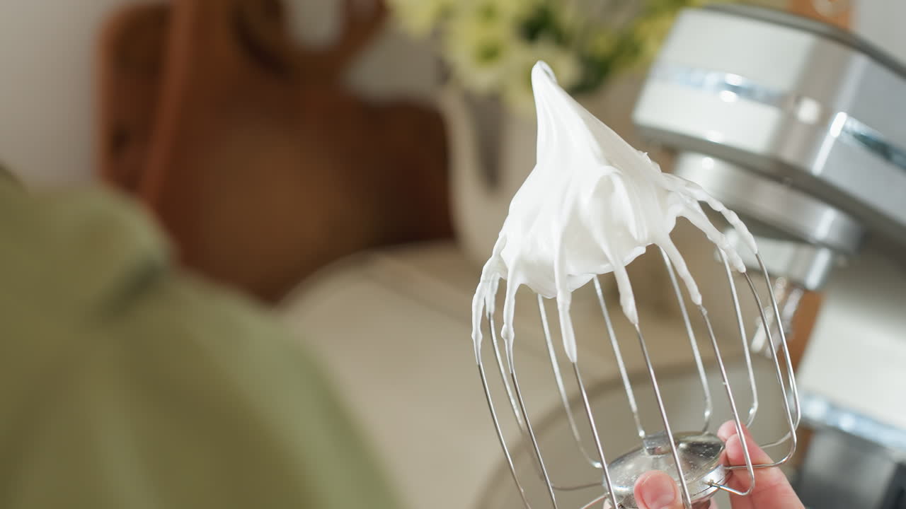 Close up of cook holding metal mixer attachment coated with thick glossy whipped meringue in kitchen setting with soft blur background, perfect for baking and dessert