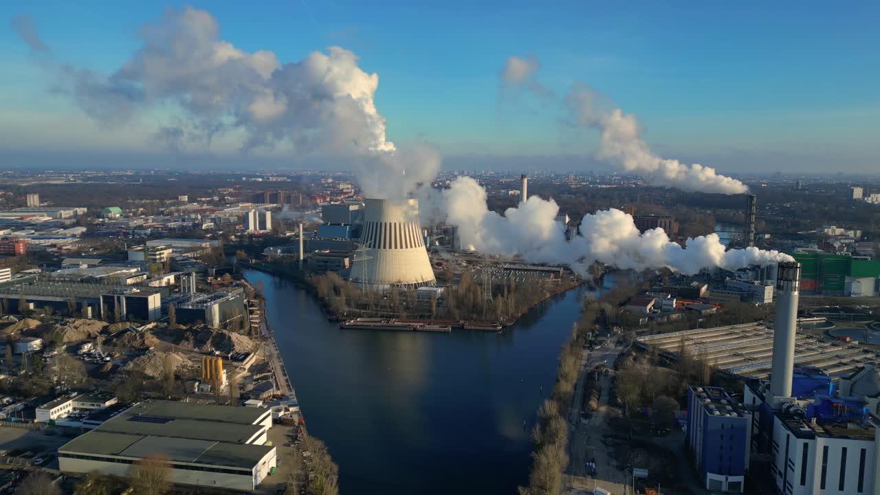 Aerial view of a thermal power plant emitting smoke on a sunny day, with a river and a city in the background. Fantastic aerial view flight