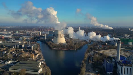 Aerial view of a thermal power plant emitting smoke on a sunny day, with a river and a city in the background. Fantastic aerial view flight