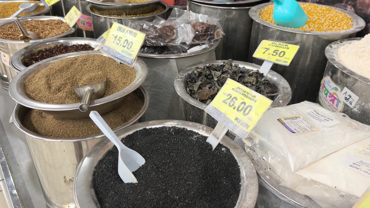 A variety of colorful spices are neatly displayed for sale in clean stainless steel jars. Each container features a clear price tag. The organized market setup looks hygienic and appealing.