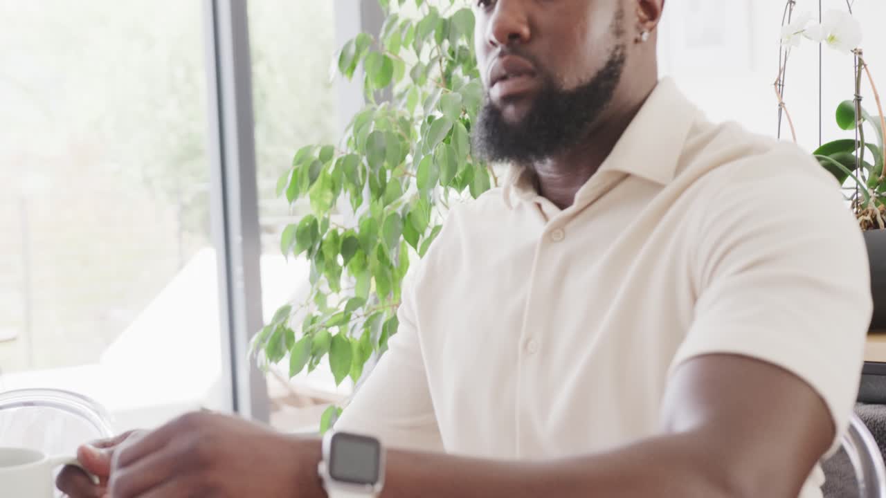 Uomo con smartwatch bianco che digita, si allunga, annusa e sorseggia da una tazza alla scrivania con il laptop.