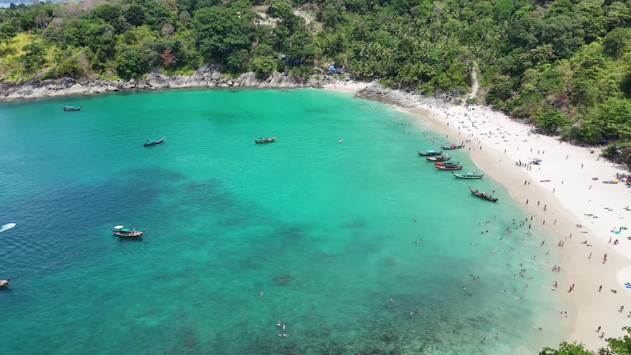las imágenes aéreas capturan una vibrante playa en phuket, tailandia, mostrando aguas turquesas, barcos y vegetación exuberante bajo la brillante luz del día.