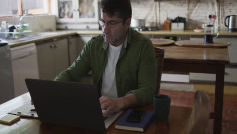 caucasian man sitting at table, using laptop and making notes at home