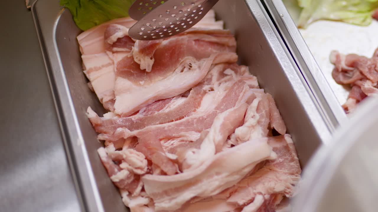 Hand uses tongs to select raw pork belly slices from stainless buffet tray under bright lighting
