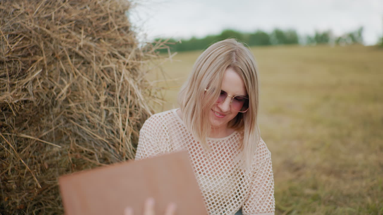 женщина в сетчатой ткани и солнцезащитных очках улыбается кому-то, держащему коричневую книгу с обложкой, с сеном на заднем плане и открытым полем, простирающимся на расстояние