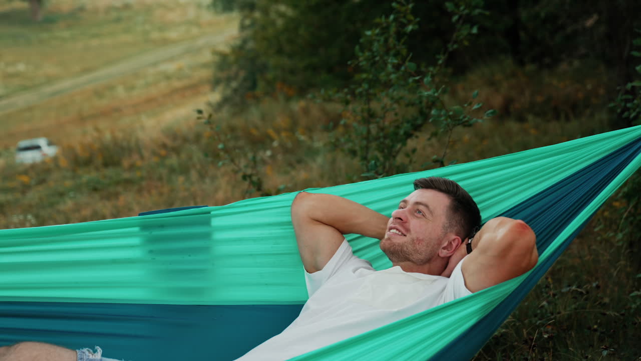 Happy relaxed freelancer working in a hammock. Man holds a laptop and types with one hand. Nature backdrop.