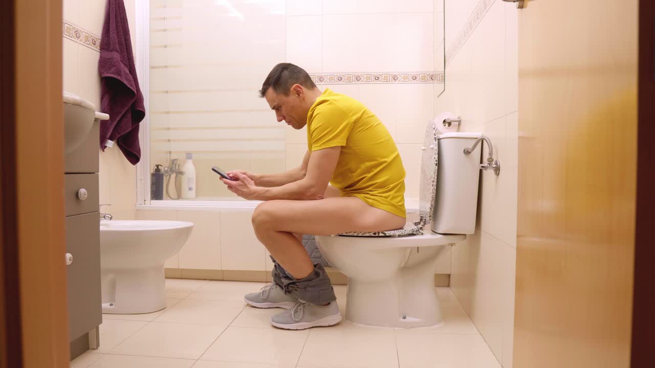Man sitting on toilet using smartphone in bathroom