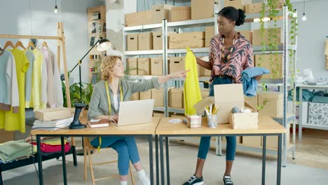 Diseñadora-Afroamericana-Empacando-Ropa-Ordenada-En-Cajas-Para-Envío-Mientras-El-Gerente-De-Ventas-Caucásico-Escribe-En-Una-Laptop-En-Una-Tienda-De-Ropa-De-Moda