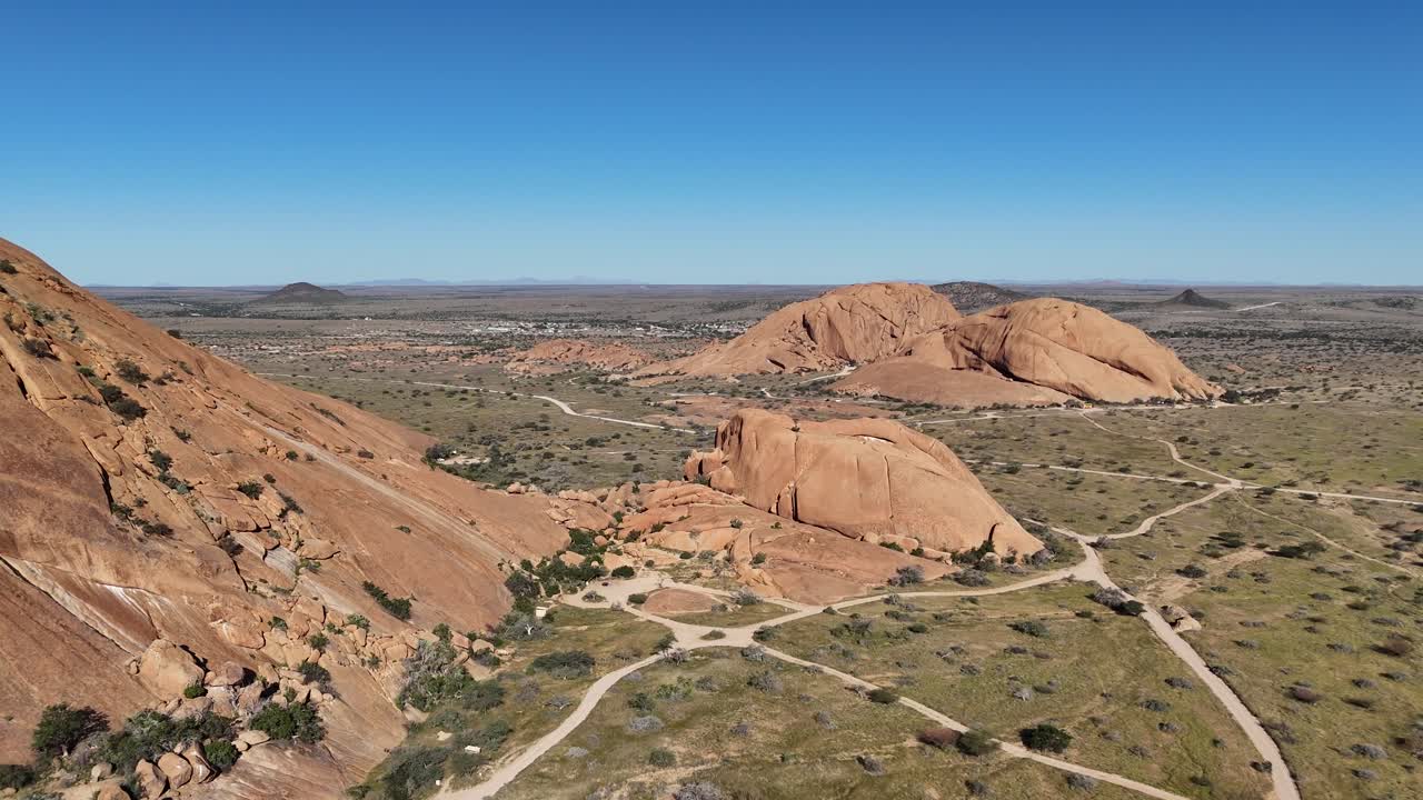 Aerial drone view of Spitzkoppe, Namibia, revealing massive granite peaks and desert trails under a clear blue sky in the Namib Desert, a famous African landmark
