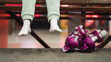 a close view of a person sitting on a bench, wearing white socks and green trousers, with pink ice skates placed beside the person, the person dangles their legs playfully