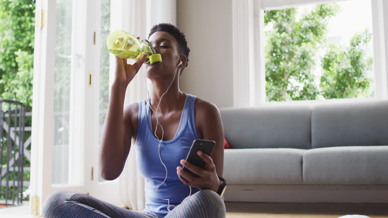 African american woman drinking water and using smartphone at home