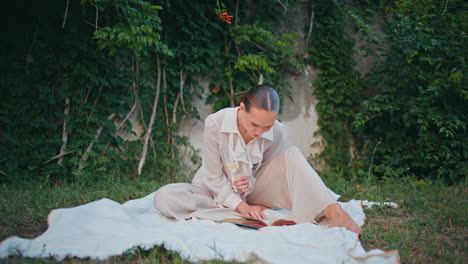 involved woman reading book on picnic blanket garden. lady holding wine glass