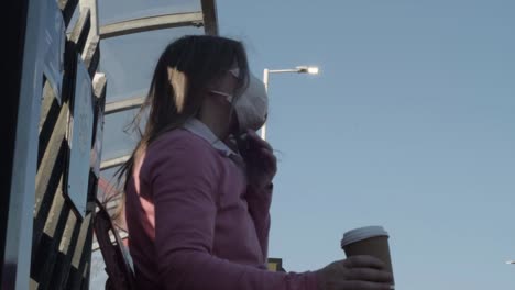 woman wearing mask drinking coffee sits down on train station bench medium shot