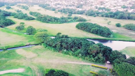 Drones-Volando-Sobre-Un-Campo-De-Golf-Sobre-Un-Lago-Y-Gente-Moviéndose-En-Los-Greens-Con-Autos-Circulando-Por-La-Carretera-Retrocediendo-Sobre-Un-Campo-De-Hierba