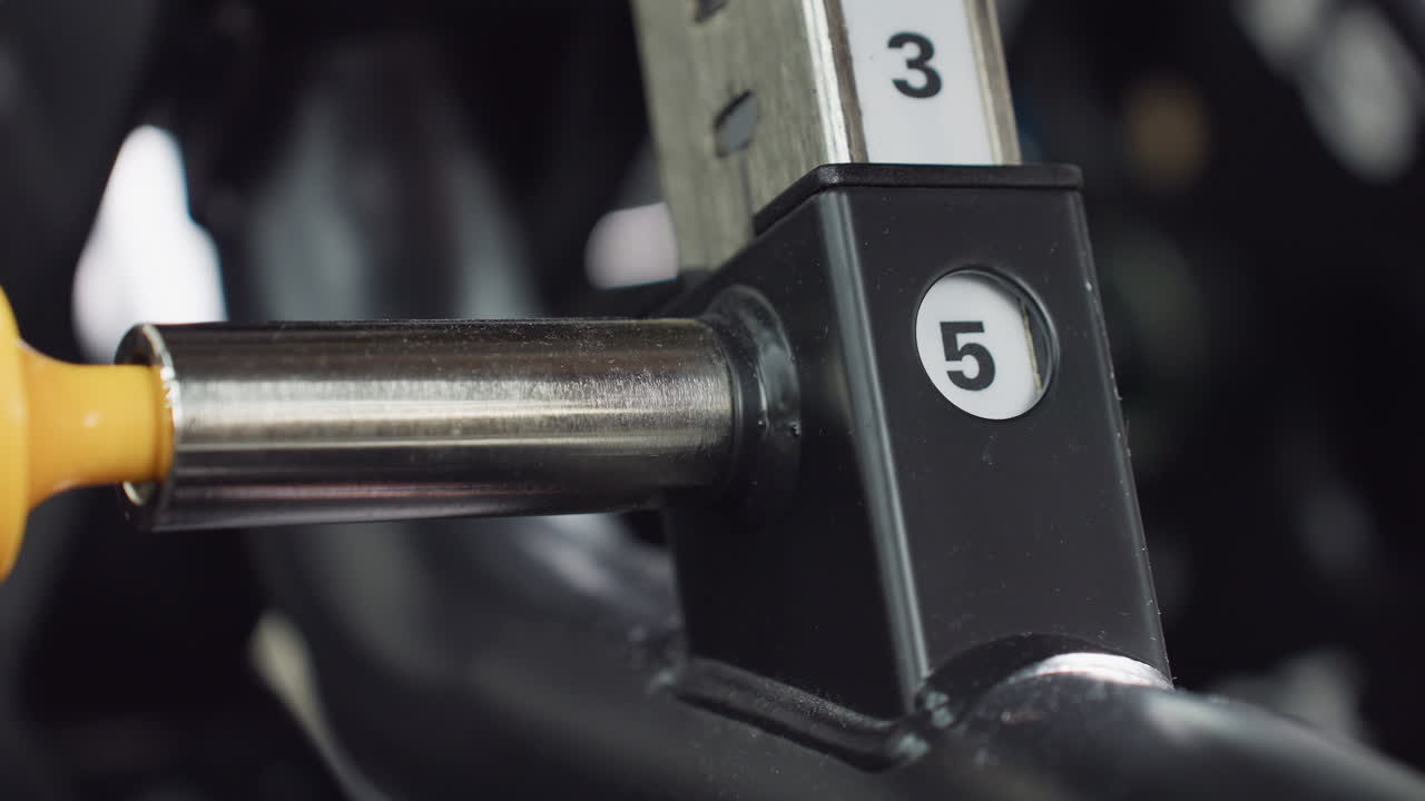 Hand pulls adjustment pin on weight bench to set seat height to five, showcasing numbered guide and metal lever in smooth motion under bright gym lighting with modern equipment detail