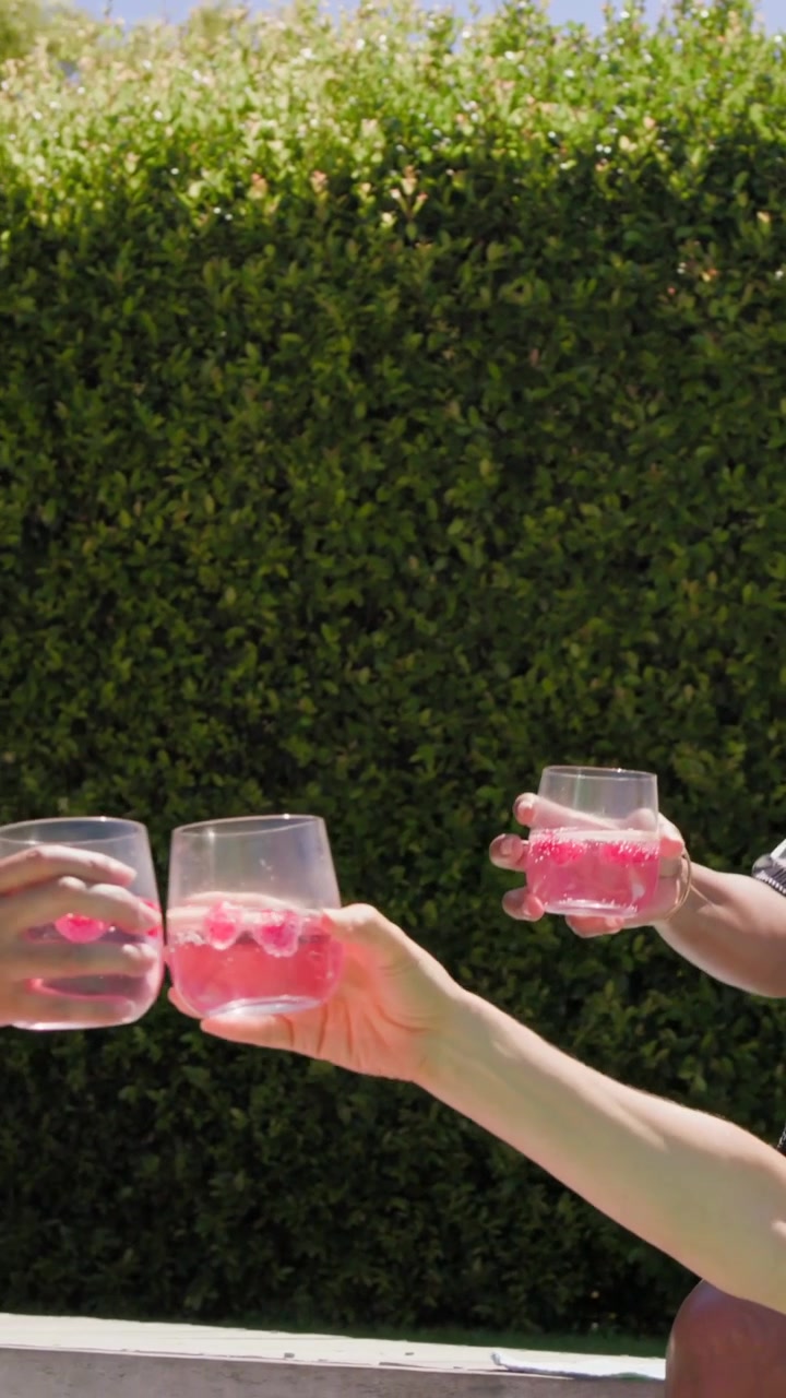Video verticale: Anziano in piscina che si allunga e porge un bicchiere rosa a una donna con copricostume e occhiali da sole, brindando.