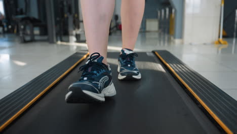 close up leg view of athlete walking on treadmill with one foot raised mid-stride, wearing canvas sneakers, gym equipment and person working out slightly blurred in background under bright lighting