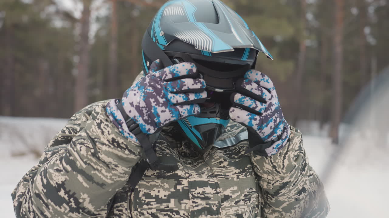 Caucasian rider adjusting helmet beside snowmobile, camo jacket and patterned gloves, mirrored goggles lowered, foggy breath in cold pine forest, methodical safety check before offroad snow ride
