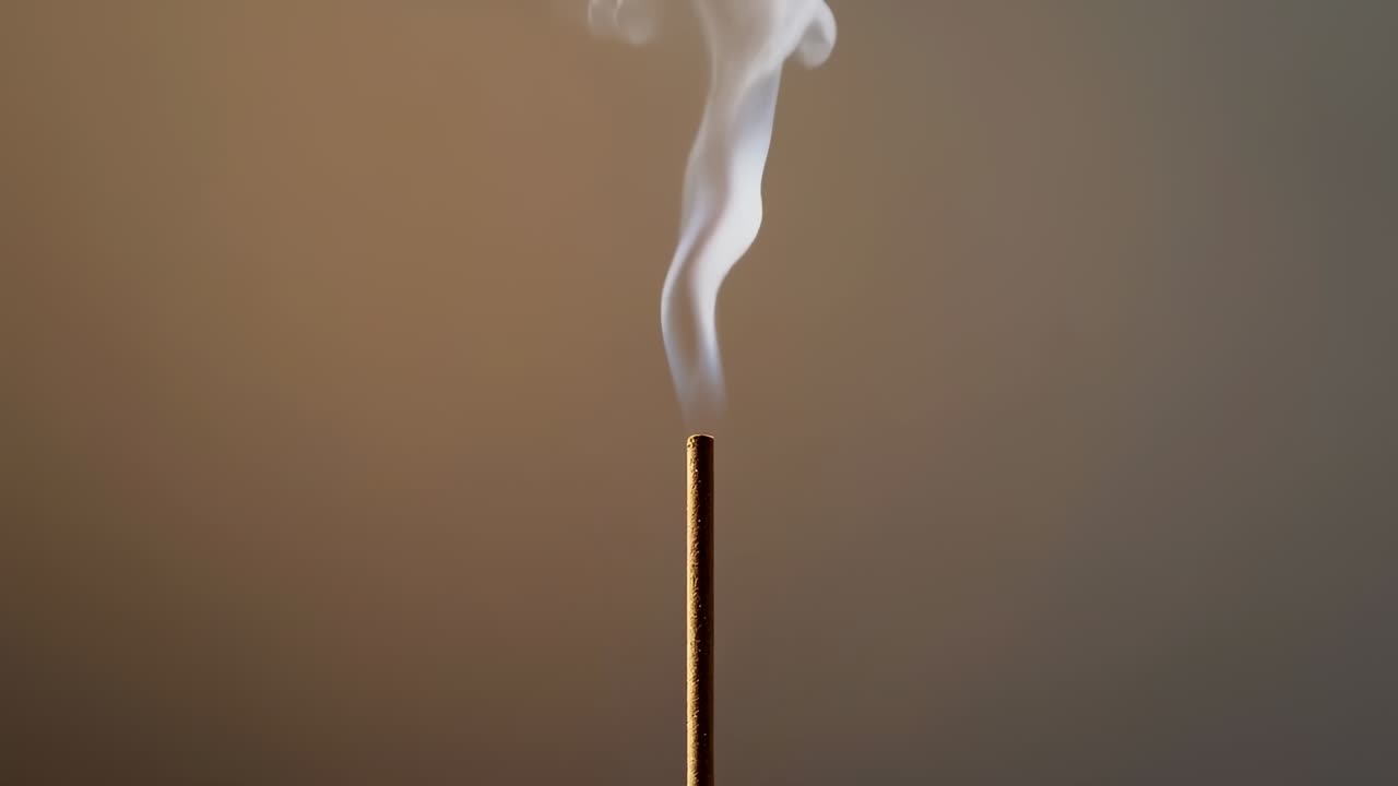 Smoldering thin incense stick releasing curling smoke in studio warm beige wall, reacting to draft