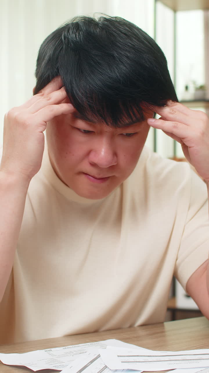 Asian man staring at due bills with calculator and laptop head in hands worried about tight budget