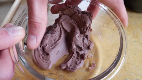 An-artist-squishing-and-shaping-very-soft,-brown-modeling-clay-in-a-glass-dish-during-a-sculpting-project-in-an-art-studio