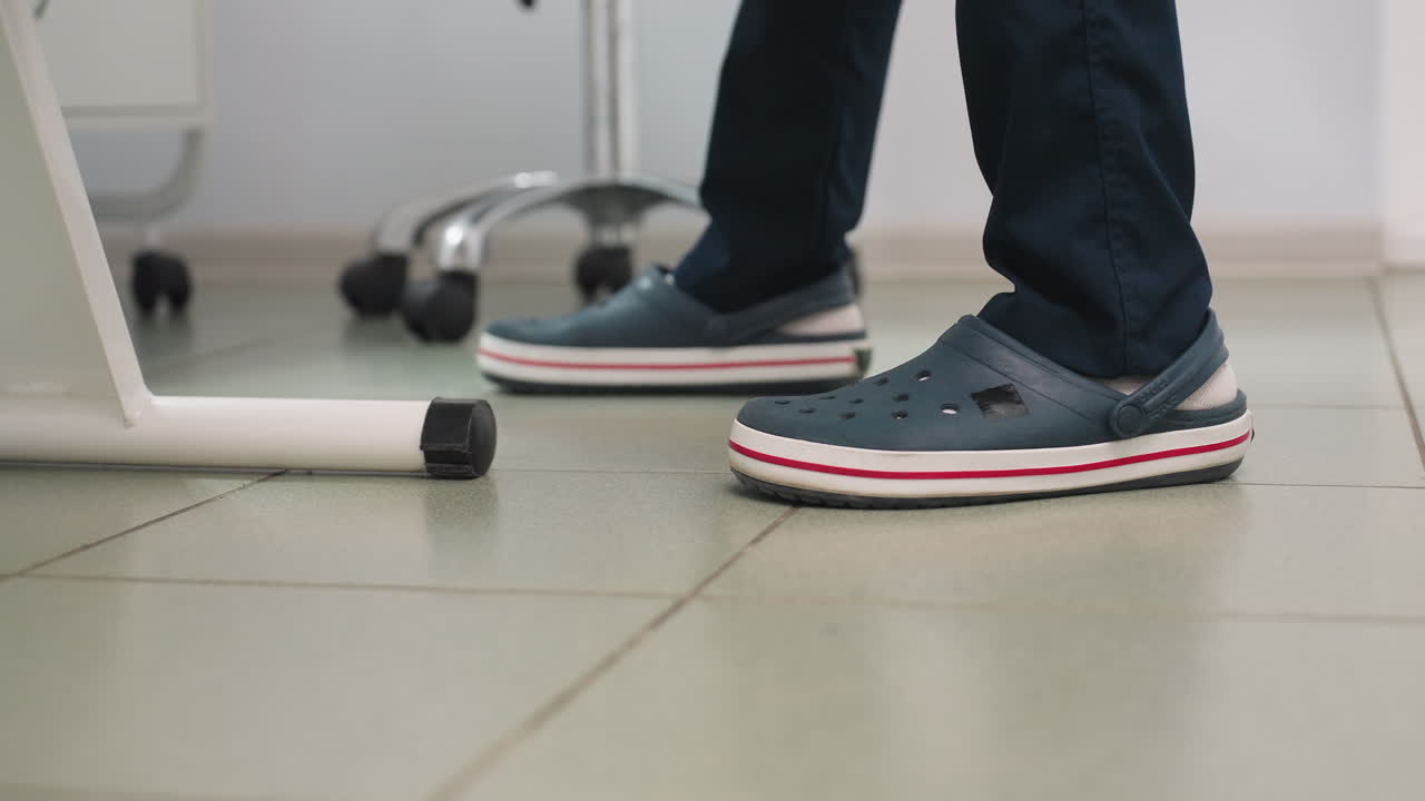 Leg view of skincare expert in Crocs with white socks standing beside massage chair on tiled spa floor with equipment trolley lamp and clean serene interior visible professional care environment