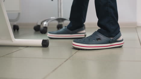 leg view of skincare expert in crocs with white socks standing beside massage chair on tiled spa floor with equipment trolley lamp and clean serene interior visible professional care environment