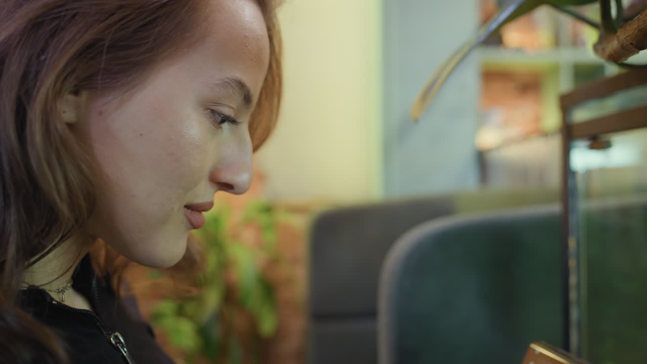 Woman with curly hair smiles softly as she captures photo of lush green environment through glass using smartphone, surrounded by cozy seating and warm interior ambiance in stylish location