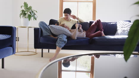 Feliz-Pareja-De-Lesbianas-Caucásicas-Tumbadas-En-El-Sofá,-Usando-Tabletas-Y-Teléfonos-Inteligentes-Y-Sonriendo-En-Una-Casa-Soleada