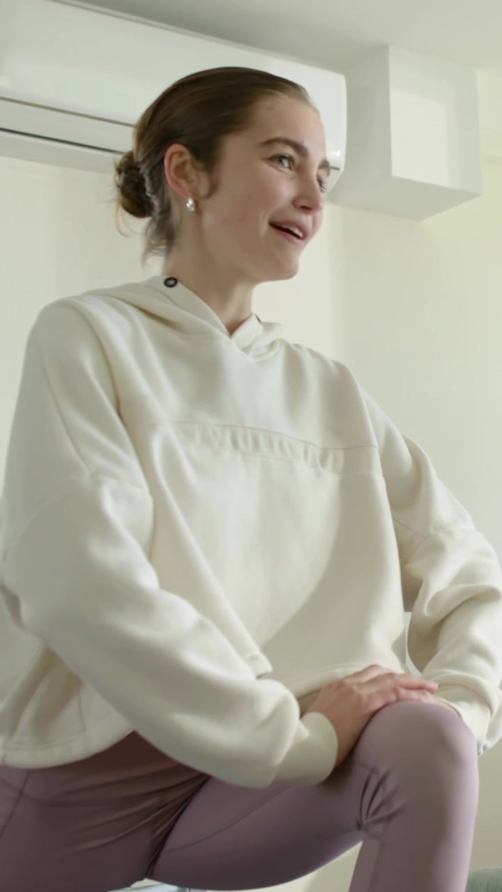 Vertikales Video: Lächelnde Frau im Ausfallschritt dreht zu Hause den Kopf nach rechts nach Geräusch außerhalb des Bildes, Kapuzenpullover, Klimaanlage.