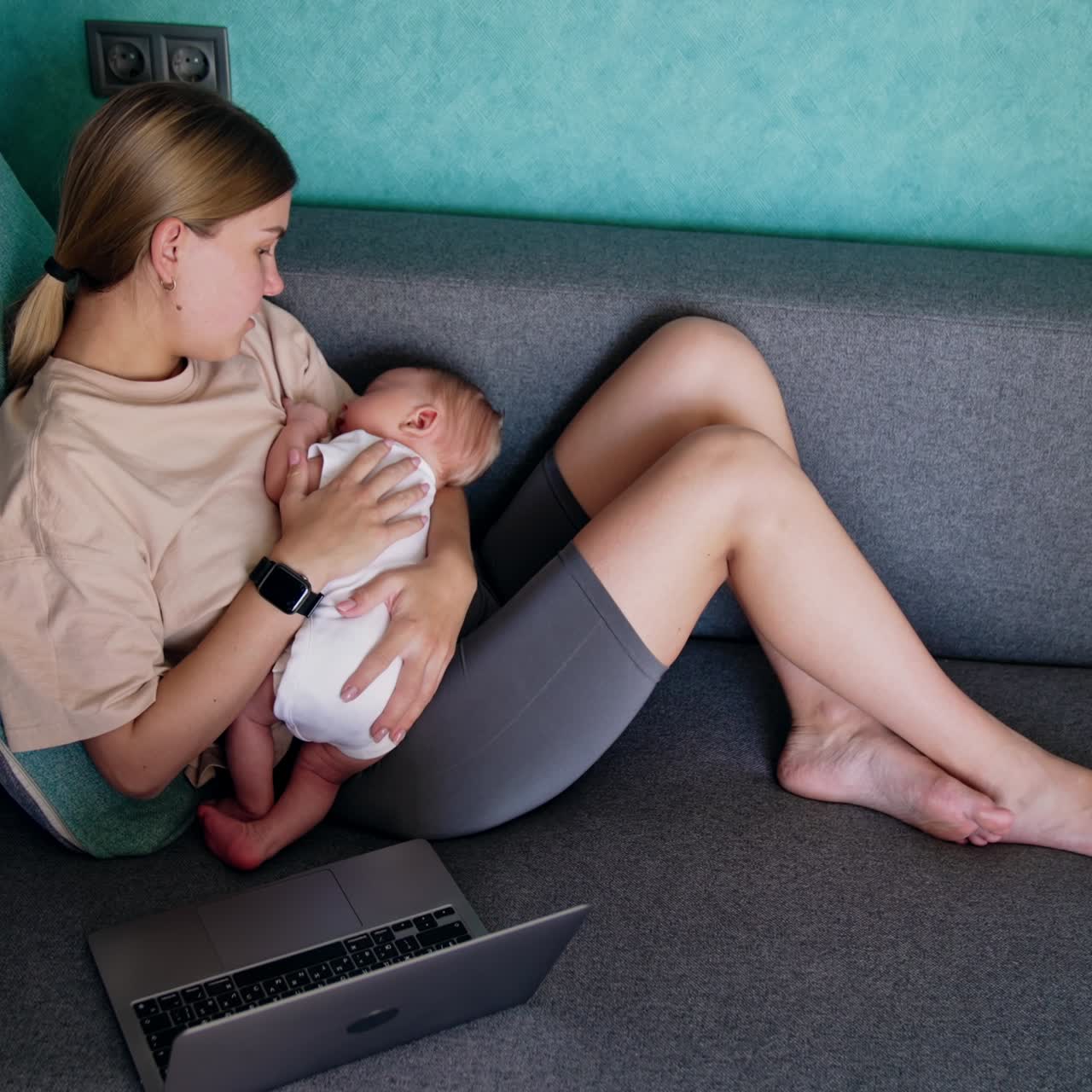 Blonde Caucasian woman with a newborn in hands sits on sofa. Opened laptop is near the lady working from home. Top view
