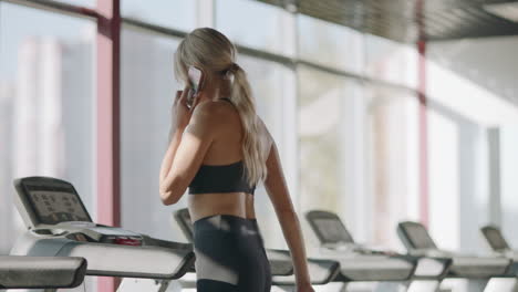 Fitness-woman-walking-on-treadmill-in-gym-club.-Pretty-girl-calling-mobile-phone