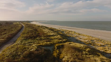 long aerial shot flying along a path through grassy dunes along an empty beach at sunset