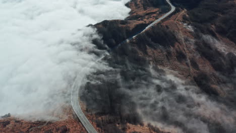 a winding road cutting through a misty mountain landscape, clouds hovering close to the ground, aerial view