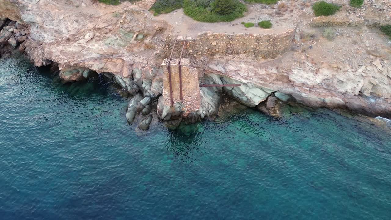 Ege Denizi'nin berrak turkuaz sularına bakan, Sifnos, Yunanistan'ın kayalık kıyısında bulunan tarihi terk edilmiş maden yükleme rampasını ve taş kalıntılarını gösteren sinematik drone çekimi