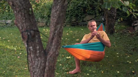 young man laying down in a hammock tied between two trees, plucking an apple out of the tree and eating it