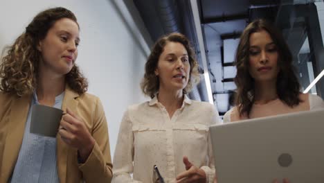 Diverse-female-colleagues-in-discussion-using-laptop-in-office-corridor,-slow-motion