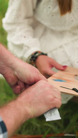 un primer plano que captura un momento de colaboración en la creación artística. una mujer sostiene una paleta de madera llena de colores vibrantes mientras un hombre abre cuidadosamente un tubo de pintura