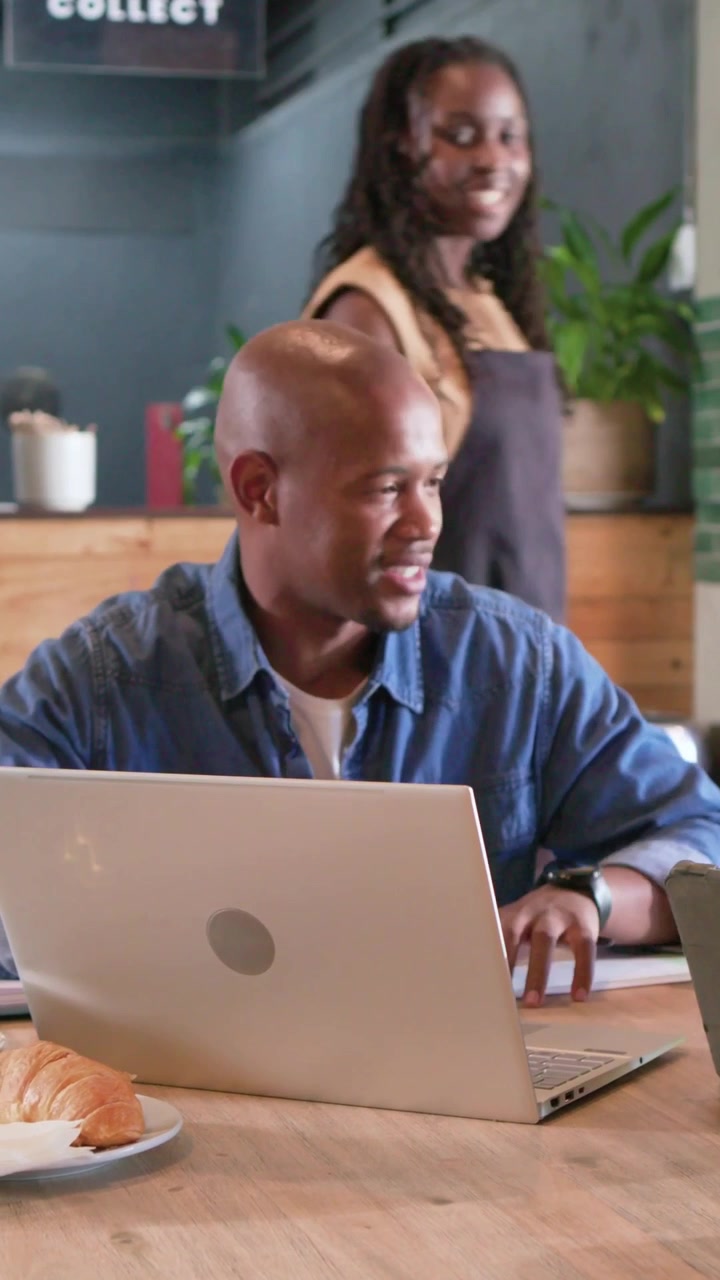 Vertical video: Noticing woman, man in denim turning right, typing and smiling at cafe with laptop