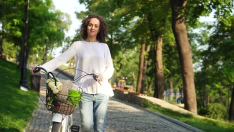Young-woman-in-a-white-t-shirt-and-blue-jeans-walking-holding-her-city-bicycle-handlebar-with-flowers-in-its-basket-in-the