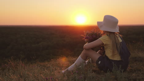 una chica con sombrero admira la puesta de sol en un lugar pintoresco, se sienta en una colina con la barbilla en las manos b