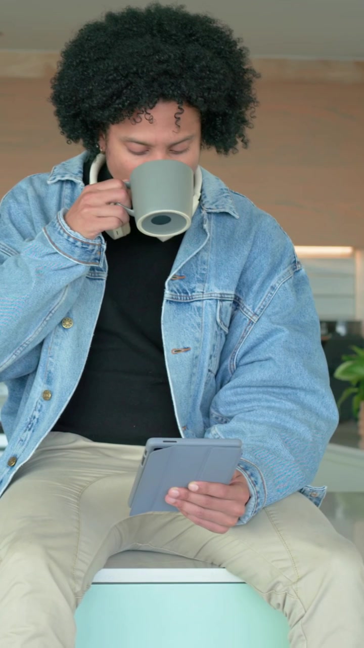Vertical video: Sitting man in denim jacket working and tapping tablet at kitchen bar, sipping mug