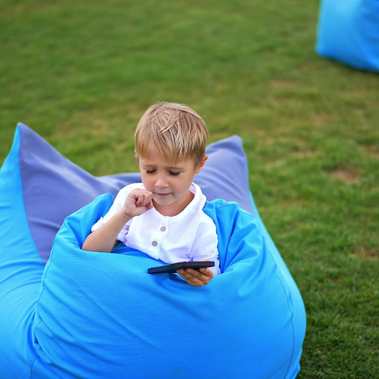 Blond preschooler having rest in bean bag chair outside. Cute boy holding phone in hand but looks aside. Circle footage