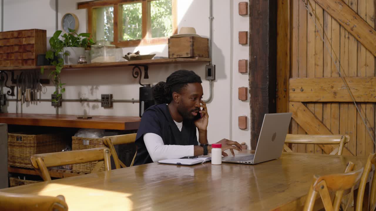 Homme afro-américain au téléphone, tapant sur un ordinateur portable, vérifiant des notes pour le travail à une table rustique, espace pour texte.