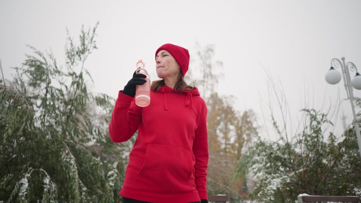 Woman drinks water from pink bottle outdoors, lowering it while gazing into distance in serene winter setting with misty atmosphere, snowy trees, and vintage streetlamp in background